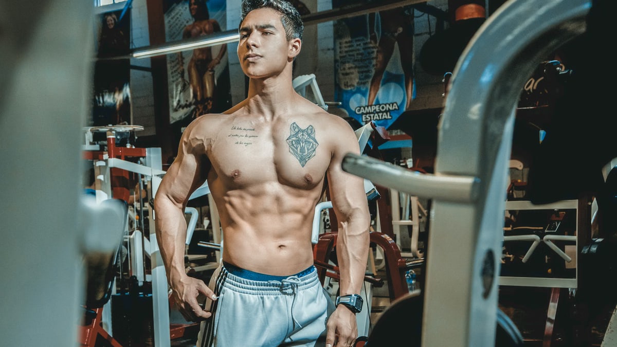 Shirtless man with tattoos posing in a gym environment, showcasing fitness and strength.